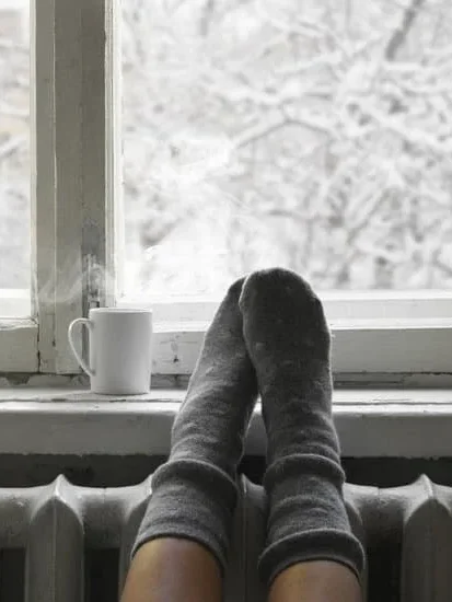 Fötter i stickade strumpor på fönsterbrädan med en kopp och snöigt landskap utanför.