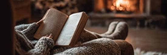En person sitter i en fåtölj och läser en bok framför en öppen eld.